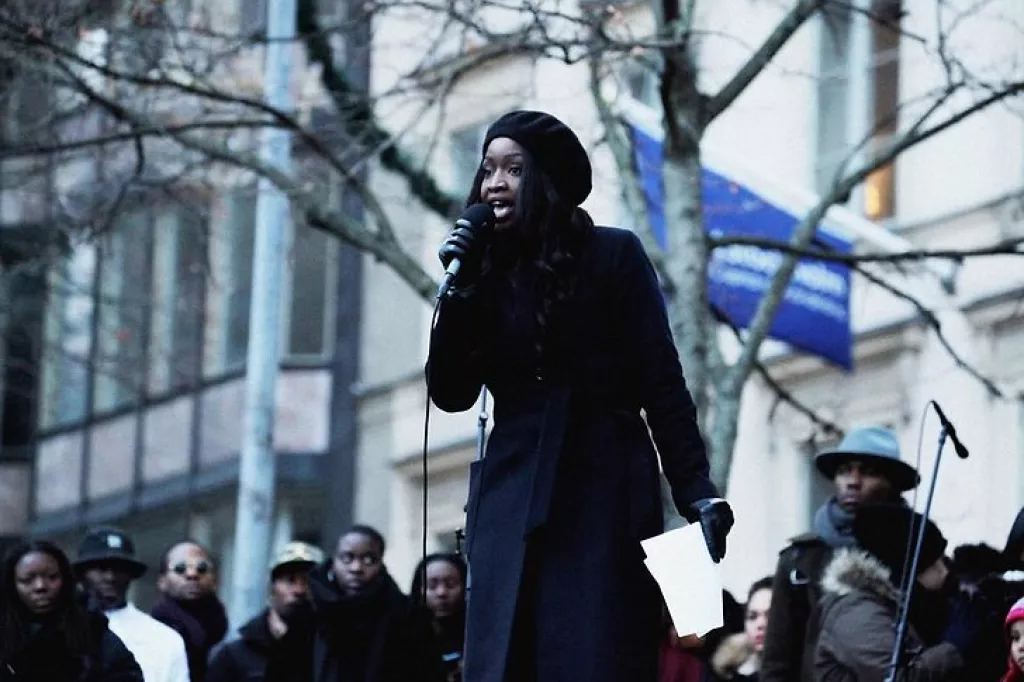 Lovette talking in a microphone in front of a big audience during cold weather. 