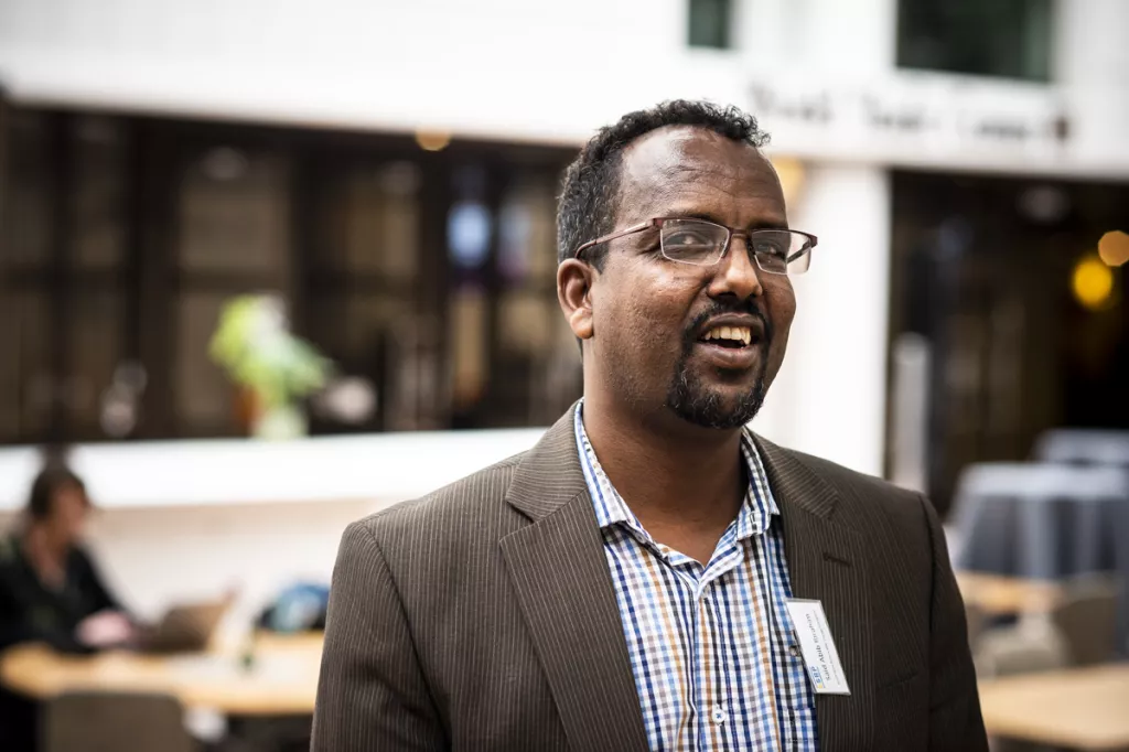 Said is smiling in a brown jacket and a checkered shirt. In the background you can see a very typical conference environment.