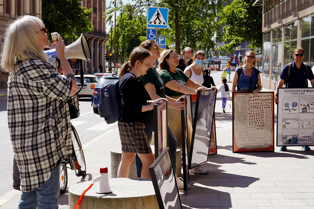 Kvinna med megafon och demonstranter håller konstverk i svarta ramar.