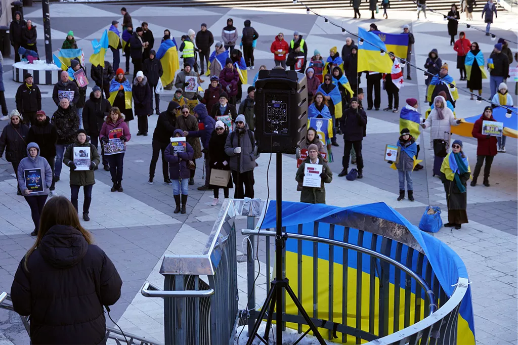 Svitlana talar framför en grupp demonstranter med plakat och ukrainska flaggor.