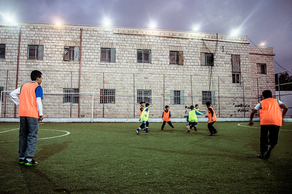 ungdomar spelar fotboll utomhus på kvällen. De har orangea och gröna västar på sig.