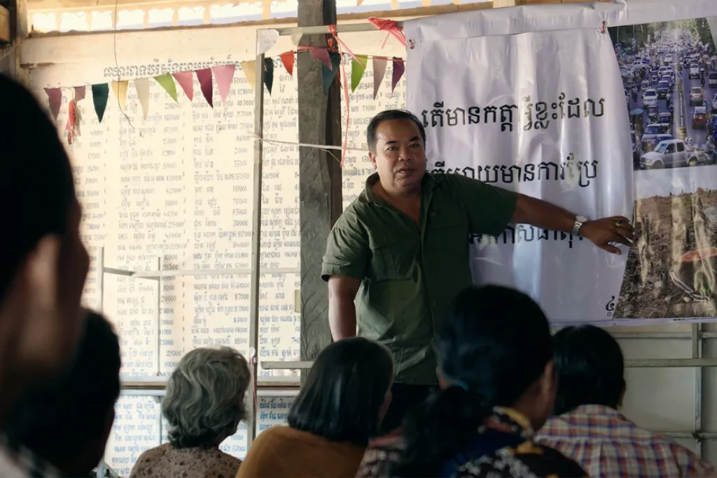 En man står framför sittande människor och förklarar bredvid en banner med text på khmer.