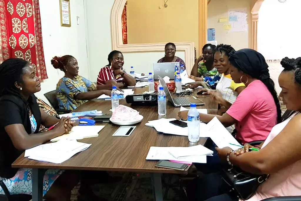 Eight women are sitting around a table with piles of papers in front of them. 