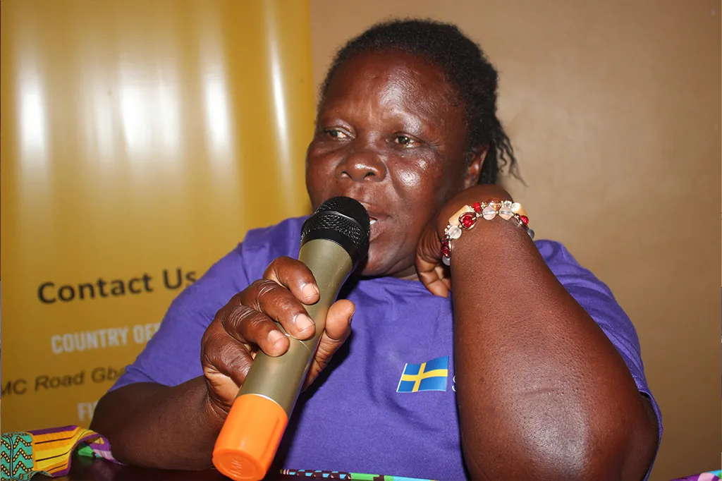 Ma Musu is wearing a purple t-shirt and is speaking into a microphone.