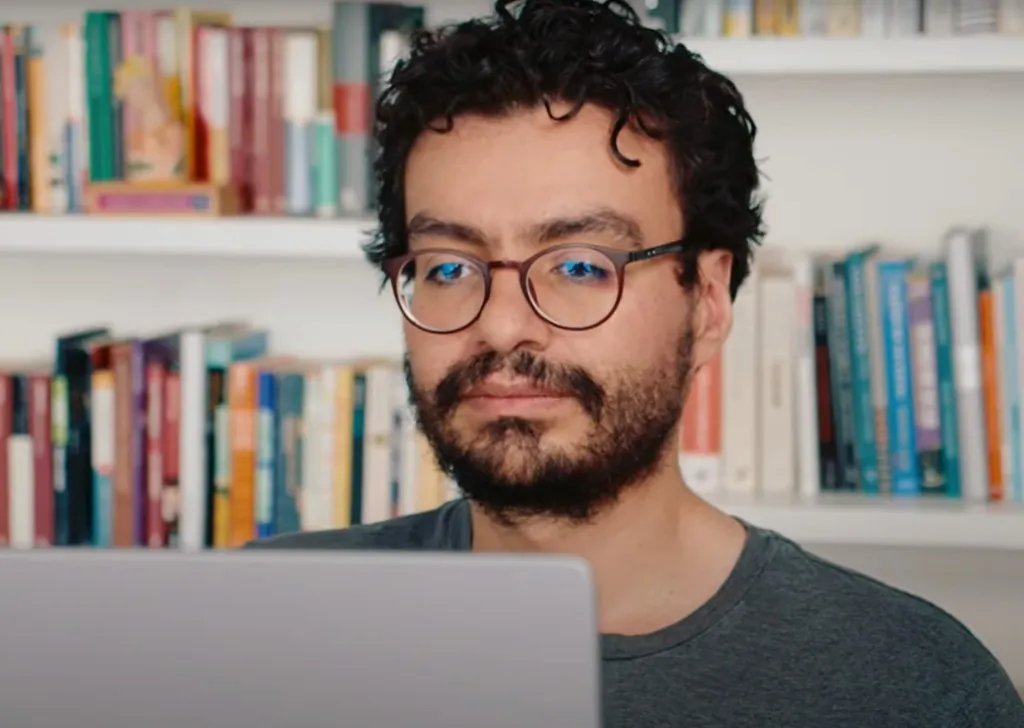 Man sitting in front of laptop with a bookshelf behind him.