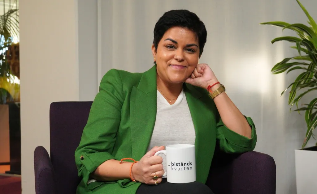 Parul iklädd grön kavaj sitter i en fåtölj