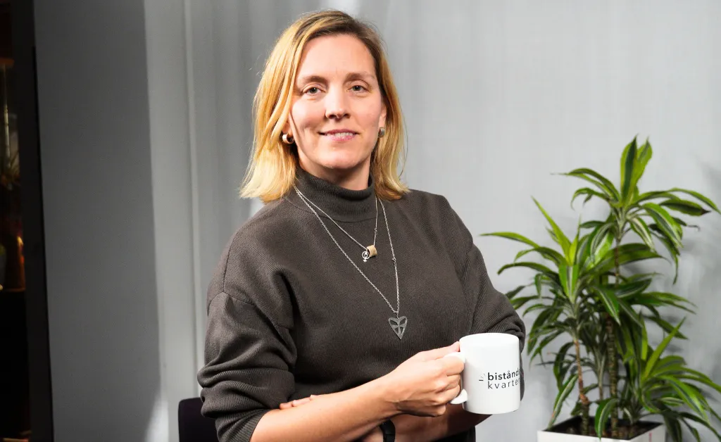 Anna håller en kaffekopp iklädd en brun polotröja