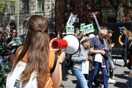 Kvinna i orange t-shirt håller i en megafon. I bakgrunden syns demonstranter med plakat.