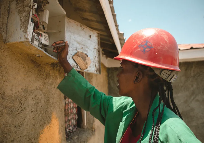 Joy is working at a electrical fuse box. She is wearing a red helmet and a green jacket.