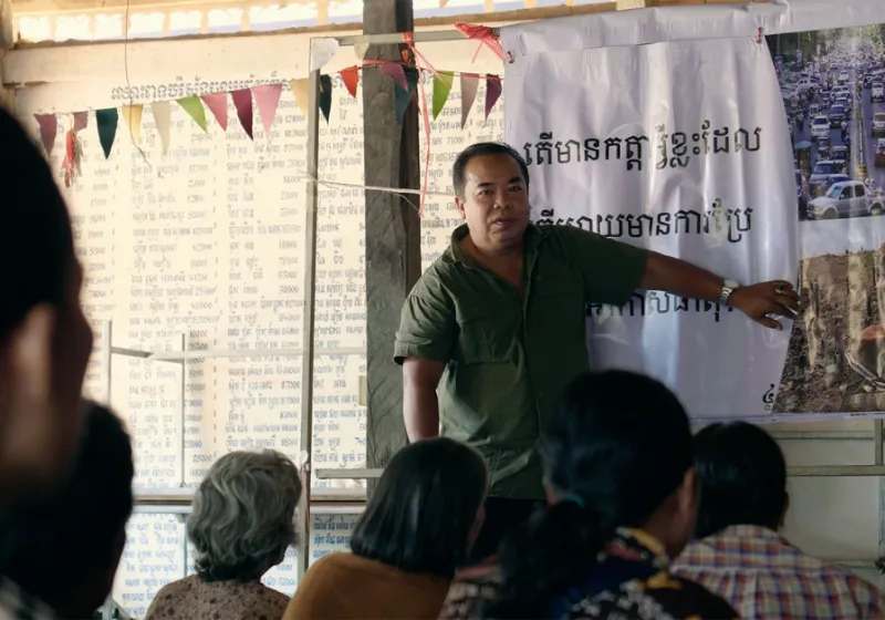 Man is standing in front of people sitting and explaining next to a banner with writing khmer