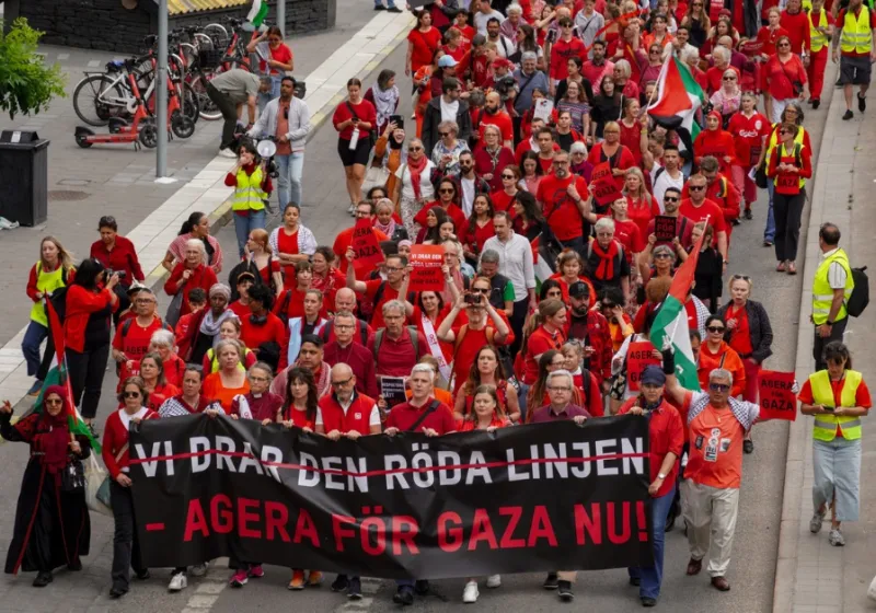 Tåg med människor klädda i rött på Sveavägen i Stockholm. Banderoll med texten vi drar den röda linjen, agera för gaza nu