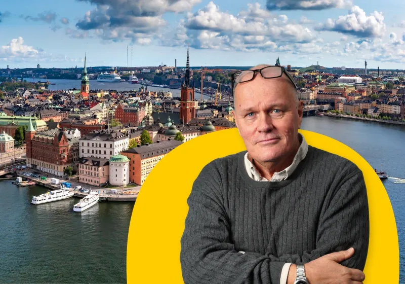 Göran i grå tröja. Bakom honom ett flygfoto över gamla stan i Stockholm. 