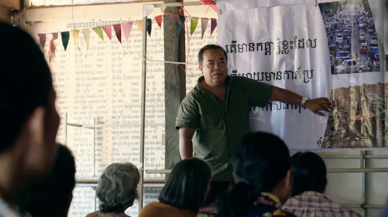 Man is standing in front of people sitting and explaining next to a banner with writing khmer