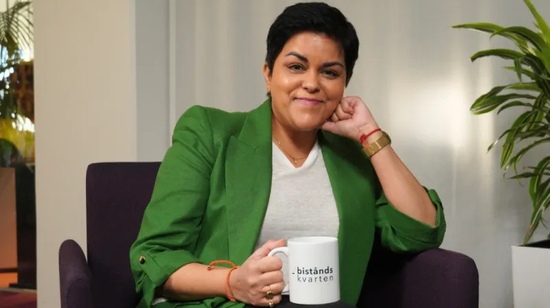 Parul iklädd grön kavaj sitter i en fåtölj