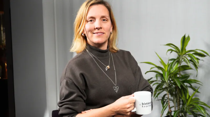 Anna håller en kaffekopp iklädd en brun polotröja